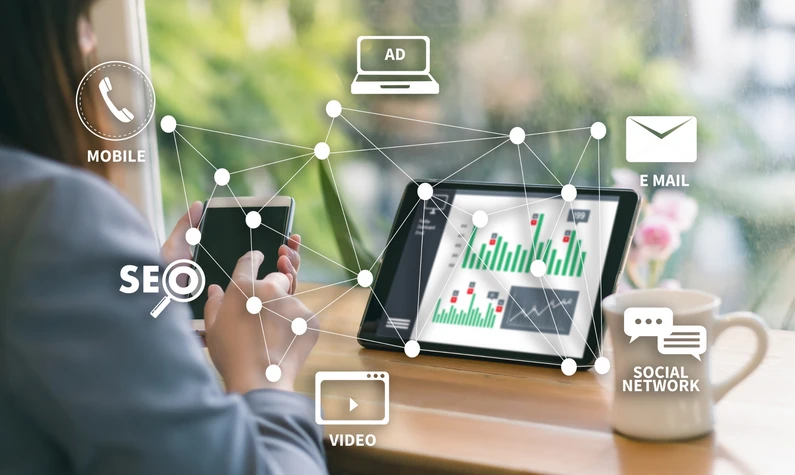 Bild som visar person med mobil framför en en tablett skärm med olika grafer som olika illustrationer som visar på en onlinemarknadsförares arbetsområden